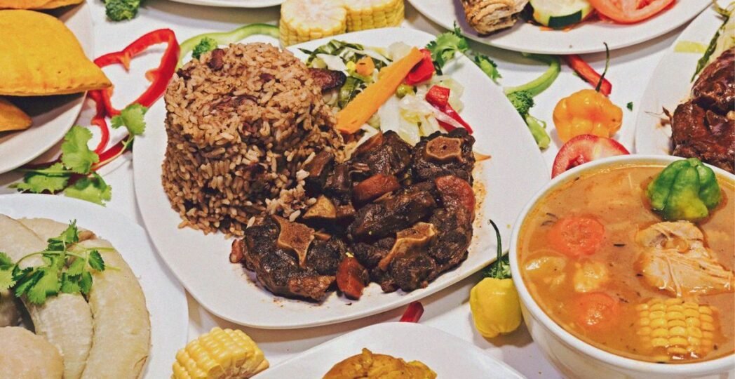 A vibrant spread of traditional Jamaican food featuring rice and peas, oxtail stew, chicken soup, and patties with vegetables and corn.