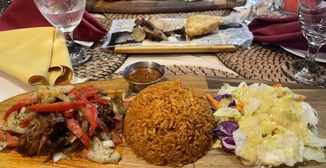A colorful plate of Kenyan food with spiced rice, grilled meat topped with onions and tomatoes, fresh salad, and dipping sauce.