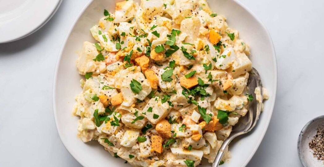 A bowl of creamy potato salad with chopped potatoes, hard-boiled eggs, celery, and fresh parsley, mixed with a mayonnaise dressing.