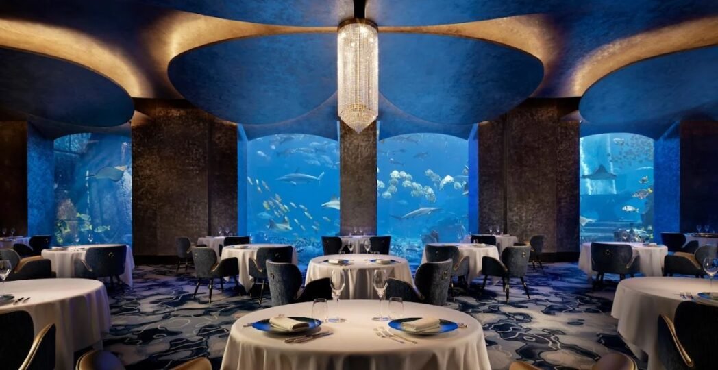 Elegant underwater restaurant with panoramic glass walls surrounded by colorful marine life — luxury dining experience beneath the ocean waves.