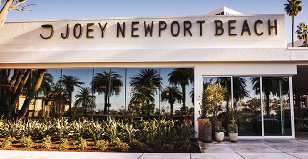 Exterior of Joey Newport Beach restaurant with large glass windows reflecting palm trees and a clean, modern white facade.